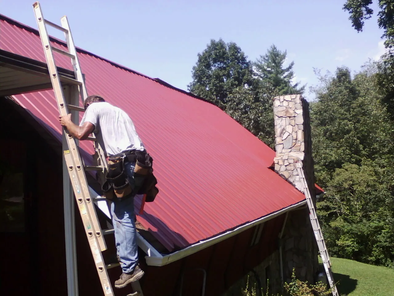 Roofer on a ladder beside a red metal roof and stone chimney — Soffit & Fascia Repair by Roofing Brothers in Pearisburg